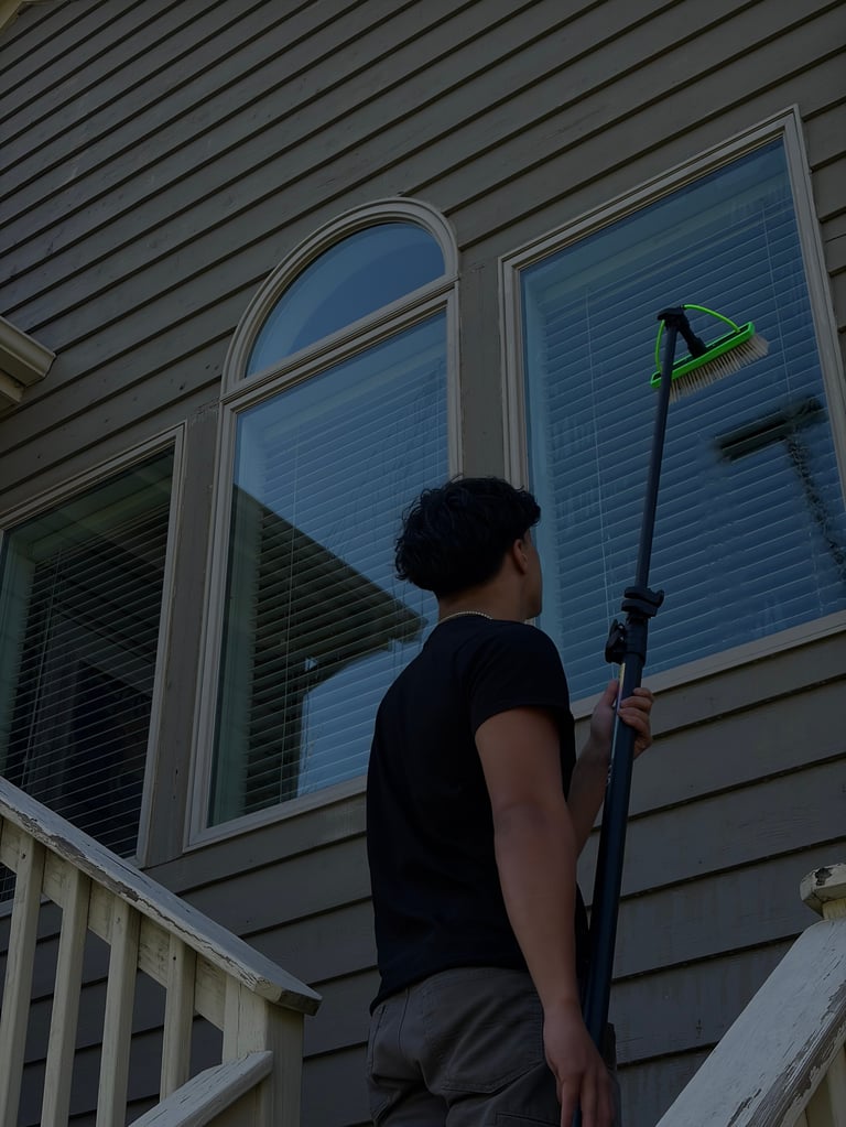 Person cleaning second-floor windows of a house using a long pole with green attachment while standing on a deck