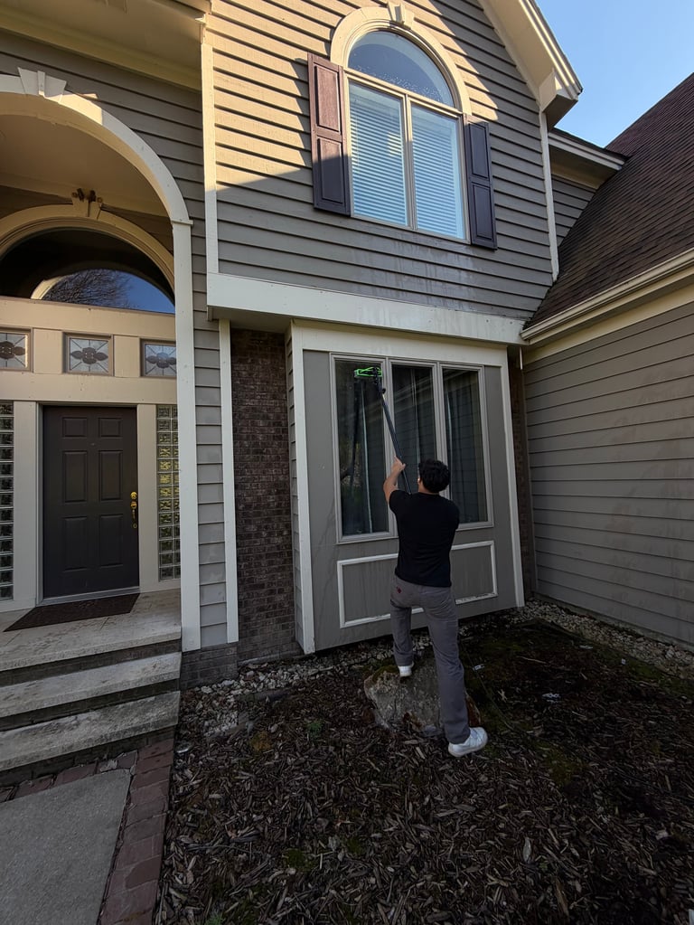 Person in black jacket cleaning windows of two-story residential house with gray siding and dark shutters